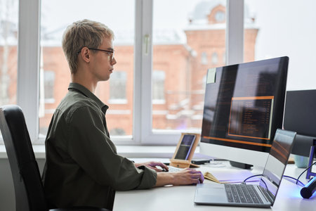 Side view of young programmer sitting at his workplace and working with security codes for program...