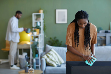African American young woman wiping dust from furniture during housework at home together with her boyfriendの写真素材