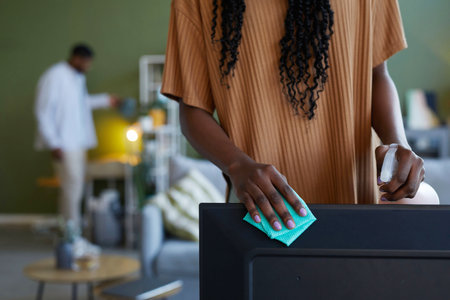 Close-up of African American housewife wiping dust from furniture while doing housework with her boyfriend in backgroundの写真素材