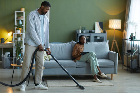 Man vacuuming the floor with vacuum cleaner in the living room, he helping his wife to do houseworkの写真素材