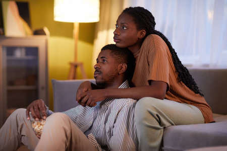 African American young couple eating popcorn and enjoying watching movie together sitting on sofa in the living roomの写真素材
