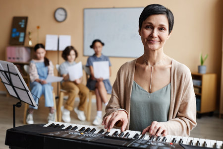 Portrait of mature music teacher smiling at camera while playing piano in music class with children in backgroundの写真素材