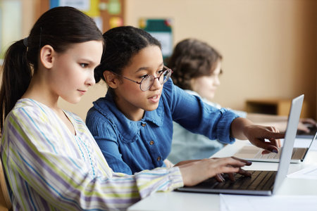 Schoolgirls sitting at table and working on laptop together during lesson in classの写真素材