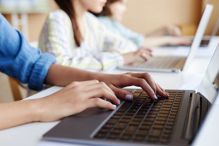 Close-up of student using laptop in study while sitting at table in class with other school children in backgroundの写真素材