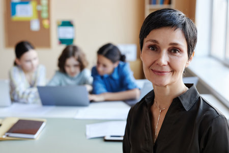 Portrait of professional teacher smiling at camera while sitting at lesson with children in backgroundの写真素材