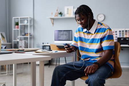 African American young man listening to music in headphones using his smartphone while sitting in officeの写真素材