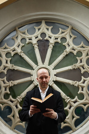 Vertical image of serious mature pastor in suit reading Bible standing against ornate in backgroundの写真素材