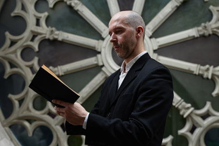 Mature man in black suit reading pray in book standing against beautiful architectural wall in churchの写真素材
