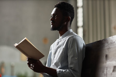 African American man sitting on bench and reading prayer from book in churchの写真素材