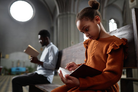 African American little girl reading Bible while sitting on bench in church with man in backgroundの写真素材