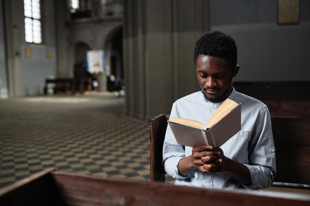African American young man sitting on bench in church and reading prayerの写真素材