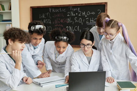 Diverse group of teen children with female teacher looking at laptop screen together in science class at schoolの写真素材