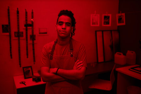 Portrait of young professional photographer looking at camera while standing with his arms crossed...