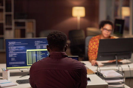 Rear view of programmer sitting at his workplace in front of computer and typing codes in office