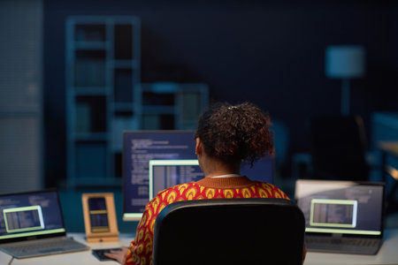 Rear view of African American woman working on computers at her workplace in IT office till late nightの写真素材