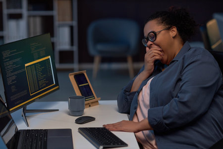 Tired programmer yawning while working at her workplace on computer till late night during deadlineの写真素材