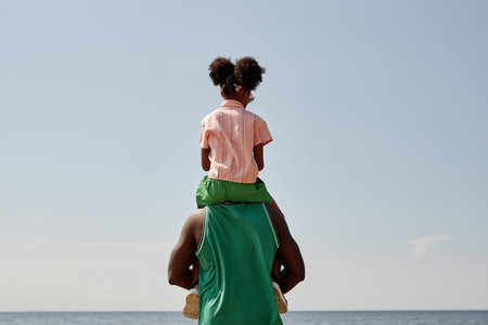 Rear view of dad standing on the beach with his little daughter on his shouldersの写真素材