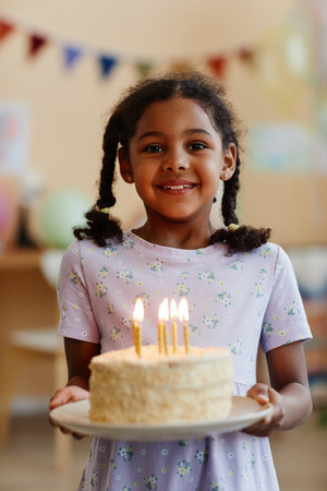 Vertical waist up portrait of black little girl holding birthday cake with candles and smiling at cameraの写真素材