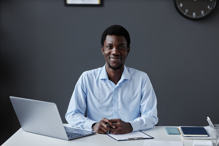 Front view portrait of young black businessman smiling at camera while sitting at workplace desk in officeの写真素材