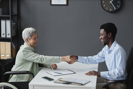 Side view portrait of smiling senior woman with disability shaking hands with recruiter after successful job interviewの写真素材