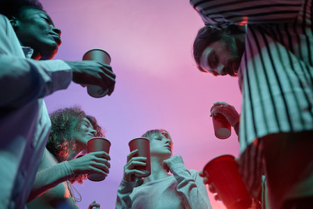 Low angle view at group of young people at house party holding red cups with alcohol and dancing in pink neon lightの写真素材
