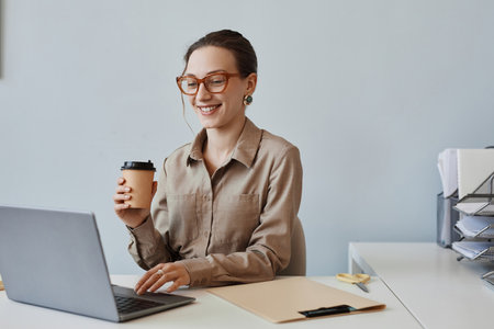 Minimal portrait of smiling young woman wearing glasses while using laptop at workplace in office...