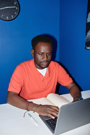 Vibrant vertical portrait of focused black man using laptop at workplace shot with flashの写真素材