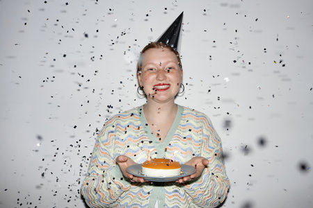 Minimal waist up portrait of smiling young woman holding Birthday cake with confetti shower at party, shot with flashの写真素材