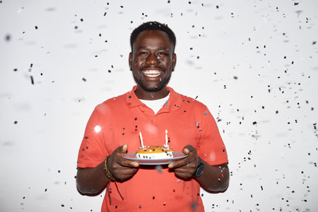 Minimal waist up portrait of smiling black man holding Birthday cake with confetti shower at party, shot with flashの写真素材