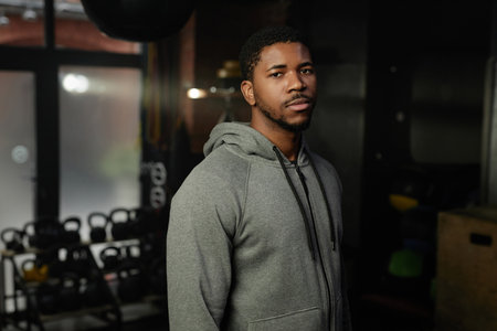 Young serious African American man in grey activewear standing in front of camera in spacious gym or...