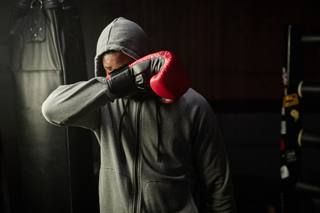 Young tired African American boxer in grey hoodie hiding his face behind red boxing glove while holding it in front of himself in sports clubの写真素材
