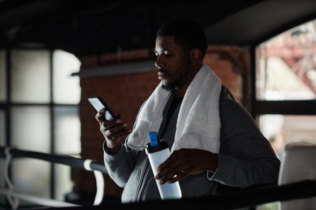 Young African American sportsman with white towel on neck having water after hard training and texting in smartphone while standing on rinkの写真素材