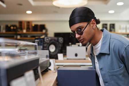 Side view portrait of black young man looking at record players and equipment in music store, copy...