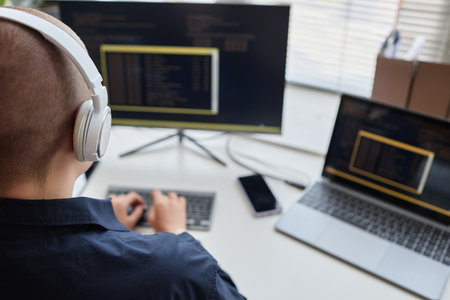 Back view of bald young woman wearing headphones and using computer while writing code and working...