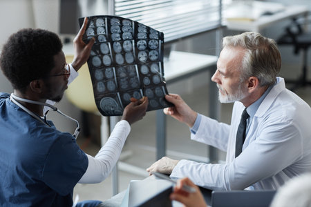 Young African American assistant showing and explaining MRI scan results of brain of patient to mature male radiologist in medical officeの写真素材