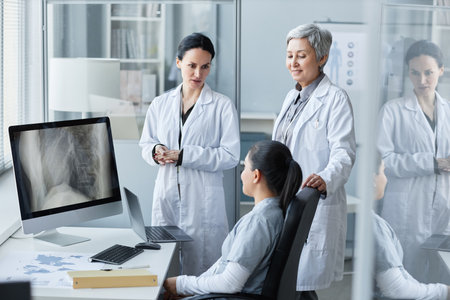 Group of three female radiologists in lab coats consulting about x-ray or MRI scan of patient on computer screen at meeting in medical officeの写真素材