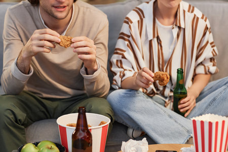 Closeup unrecognizable young couple eating fried chicken fast food while watching TV at homeの写真素材
