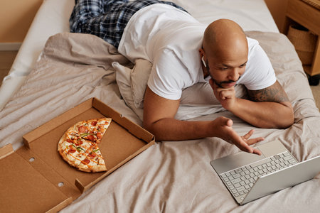 Top view of adult black man lying in bed at home and enjoying lazy weekend with pizzaの写真素材