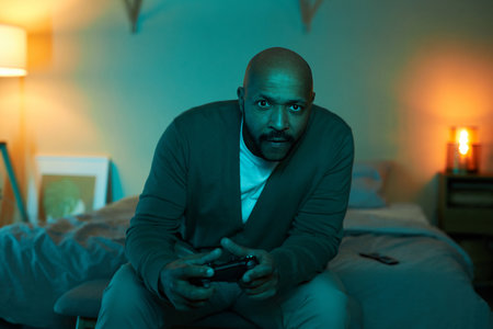 Front view portrait of adult black man playing video games at home in dark, copy space