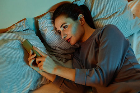 Top view portrait of smiling adult woman using smartphone in bed at night reading text messages and scrolling social mediaの写真素材