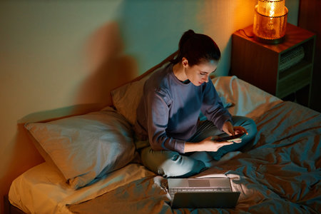 High angle portrait of young woman working late at home while sitting on bed with laptop and smartphoneの写真素材