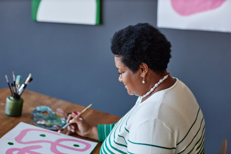 Mature female artist in casualwear painting with gouache or water colors on sheet of paper while sitting by desk in studio of arts and designの写真素材