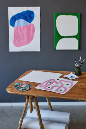 Wooden desk with paper sheets, watercolor palette and small jar with paintbrushes standing against...