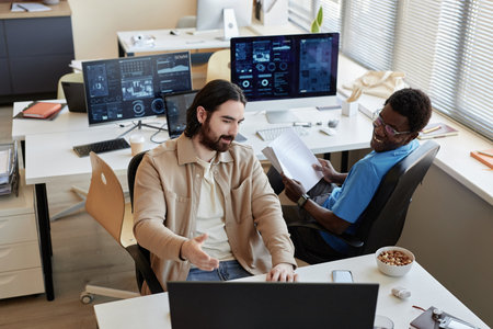 Young businessman showing data on computer screen to smiling African American colleague while...