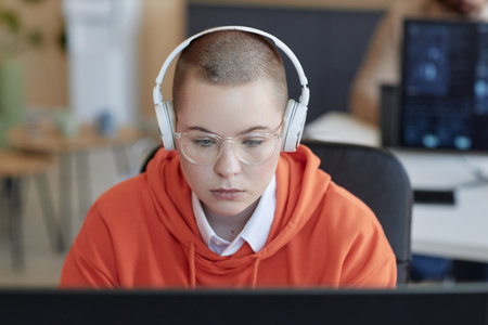 Young serious female analyst in eyeglasses and headphones looking at screen of computer and thinking...