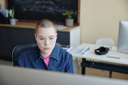 Young serious female IT manager in eyeglasses looking at computer screen while sitting by workplace...