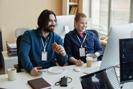 Two happy young office managers having snacks at lunch break while sitting in front of computer...