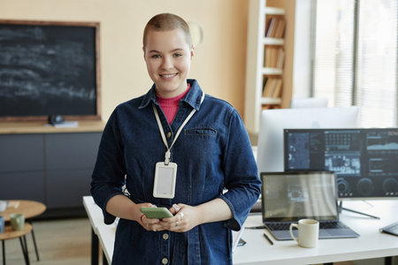 Young smiling businesswoman using mobile phone and looking at camera while standing by workplace...