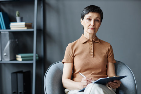 Portrait of mature psychologist sitting on armchair and looking at camera while making notes in card at sessionの写真素材