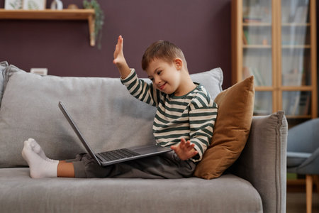 Side view portrait of happy little boy with down syndrome using laptop on sofa and waving to video...